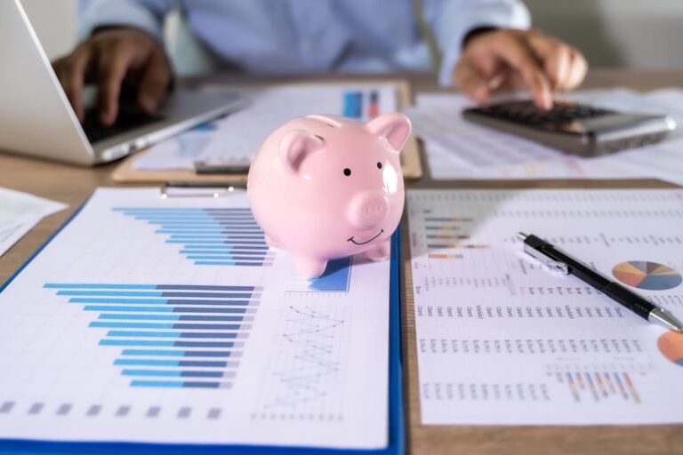 renda passiva: uma pessoa realizando cálculos com uma calculadora e algumas folhas de gráficos em primeiro plano mais um cofrinho de porquinho.
