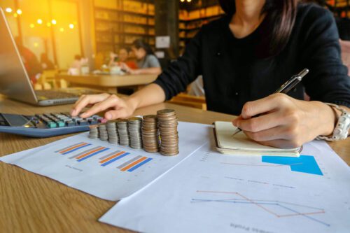 Mulher usando calculadora e anotando em caderneta. À sua frente, uma pilha de moedas em ordem crescente simbolizando a renda fixa.