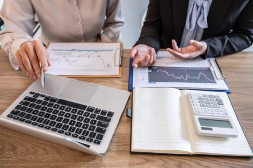 consultoria de investimentos: duas pessoas analisando o mercado financeiro, olhando no notebook e gráficos em cima da mesa.