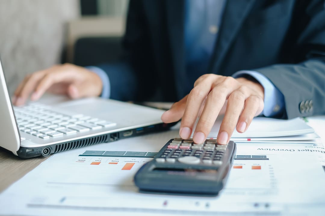 fundos de investimento em direitos creditórios: uma homem no fundo usando a calculadora e acompanhando as oscilações do mercado financeiro na tela do notebook.
