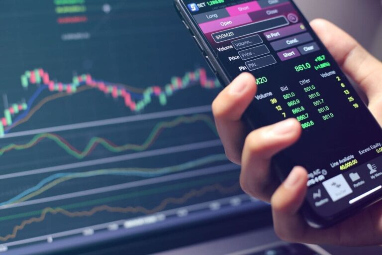 day trade: uma pessoa com celular na mão acompanhando as oscilações das ações na Bolsa de Valores e também no computador.