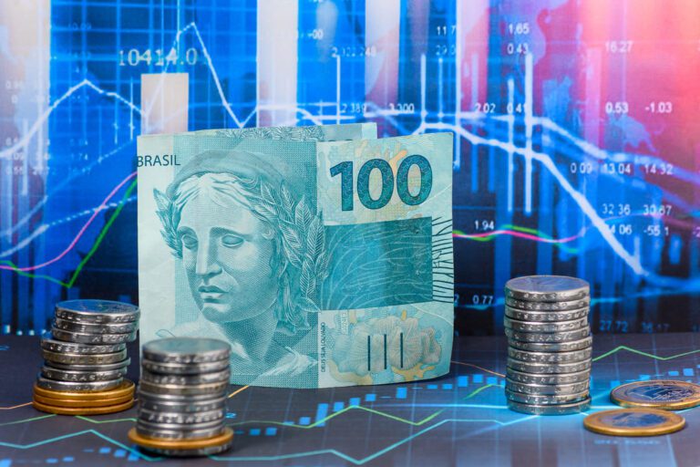 inflação no Brasil: uma nota de 100 reais e pilhas de moedas e em segundo plano um gráfico mostrando as oscilações do mercado financeiro.