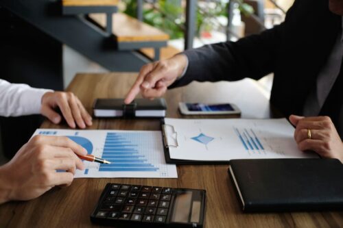 Investidor qualificado: foto de mesa de madeira marrom com papéis com gráficos, calculadora e célular, além de uma pessoa de cada lado indicando conversa