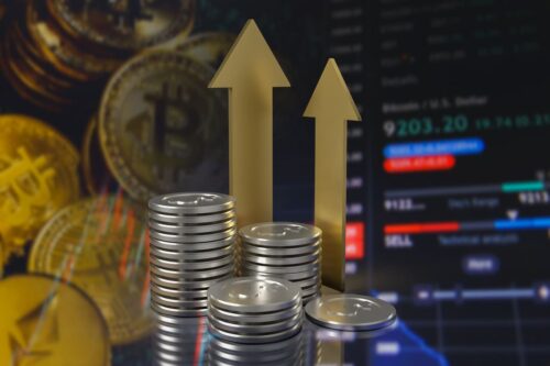 dividend yield: algumas pilhas de moedas na cor prata em primeiro plano e em segundo duas seta douradas e um monitor mostrando preço de ações.