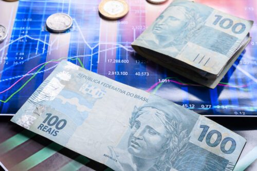 pib brasileiro: notas de 100 reais dobradas e uma aberta e moedas de um real, no segundo plano um gráfico de barra.