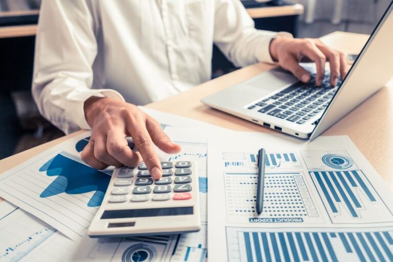 Mercado futuro de ações: pessoa branca com camisa social com uma mão na calculadora e outra no notebook em mesa cheia de papéis