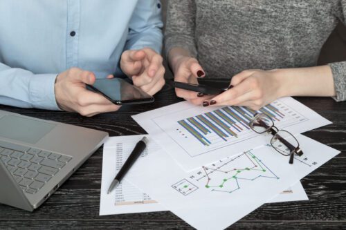 Casal mexendo no celular e analisando folhas de gráficos e notebook, representando o relatório do balanço patrimonial. Uma caneta e óculos estão sobre os papéis.