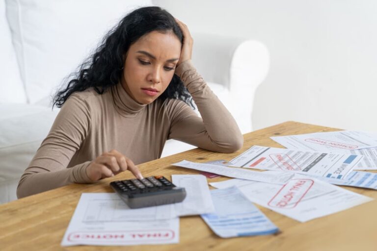 Mulher sentada usando calculadora e com expressão de preocupada olhando para vários papéis, pensando em como sair das dívidas.