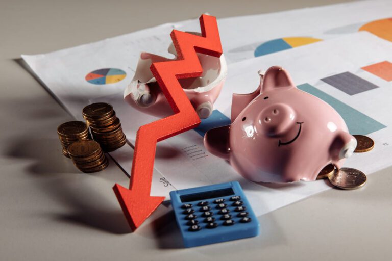 Superfície com cofre de porquinho quebrado sobre papéis, moedas empilhadas, calculadora azul e seta vermelha apontada para baixo, indicando risco de liquidez.