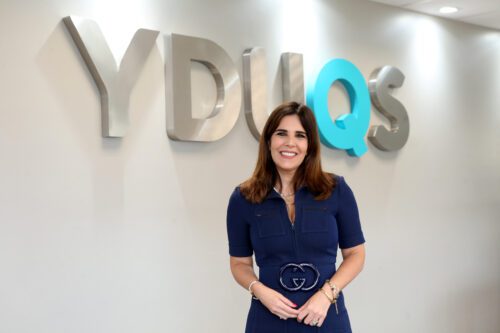 logo da Yduqs em prata destacando o q em azul turquesa em uma parede simples com uma mulher na frente