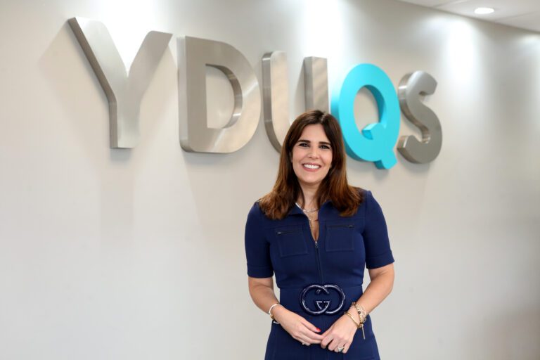 logo da Yduqs em prata destacando o q em azul turquesa em uma parede simples com uma mulher na frente