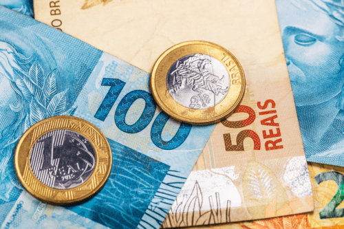 Notas de 100 e 50 reais, e moedas de 1 real espalhadas. A divisa surgiu do chamado Plano Real.