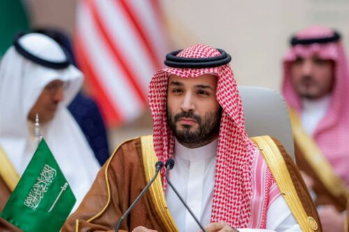 Mohammed bin Salman, o príncipe herdeiro da Arábia Saudita, em uma reunião, com figuras desfocadas ao fundo e bandeiras ao redor.