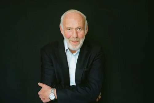 Retrato de Jim Simons, fundador da Renaissance Technologies, com fundo preto, usando terno preto e relógio prateado.