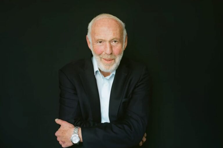 Retrato de Jim Simons, fundador da Renaissance Technologies, com fundo preto, usando terno preto e relógio prateado.