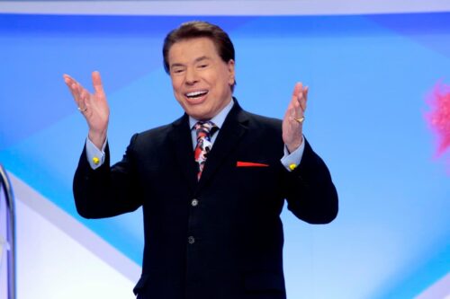 Silvio Santos sorrindo e gesticulando com as mãos durante a gravação de seu programa de televisão em estúdio.