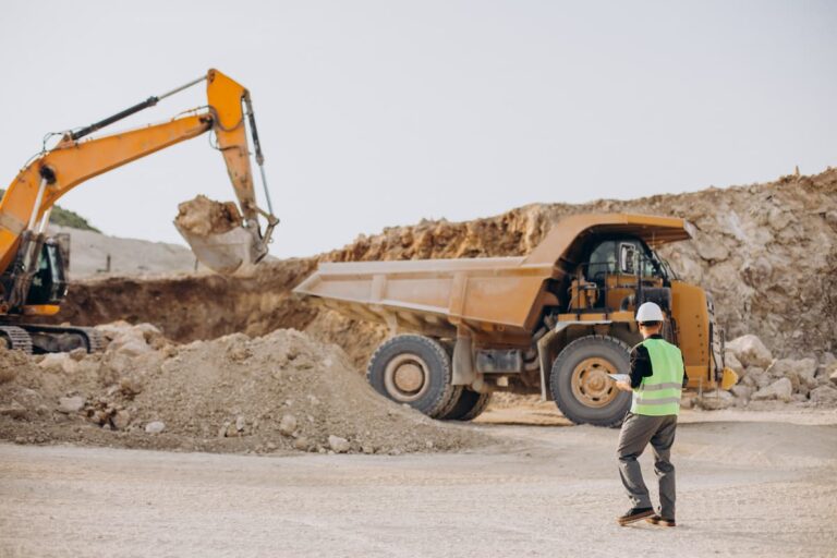 Homem com colete de segurança observa escavadeira carregando caminhão de minério em mina a céu aberto, representando empresas de mineração na Bolsa de Valores.