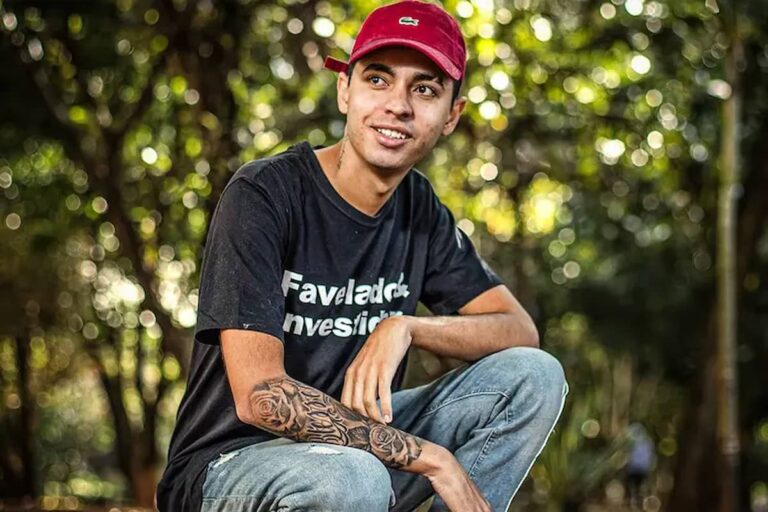 Murilo Duarte, sorridente, usa boné vermelho, camiseta preta com a inscrição "Favelado Investidor", sentado ao ar livre em ambiente arborizado.