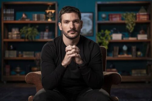 Foto de Bruno Perini sentado, de mãos cruzadas, em uma sala com estantes ao fundo, vestindo uma blusa preta.