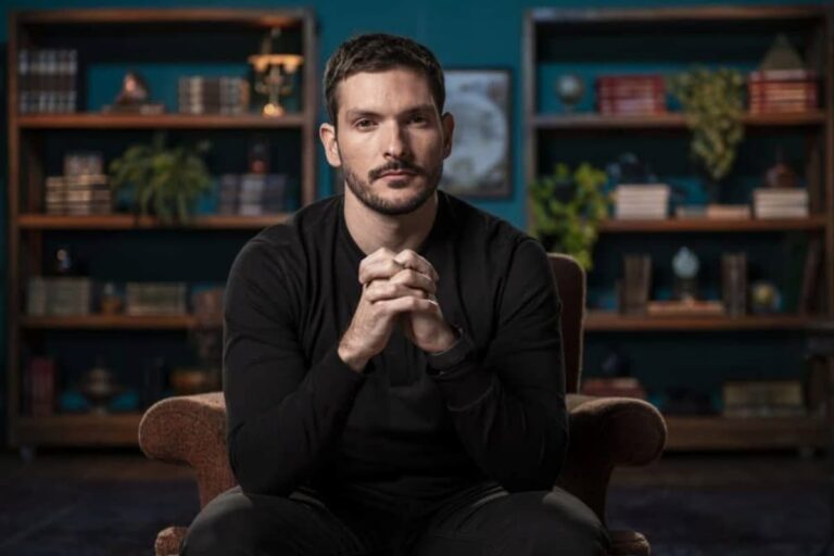 Foto de Bruno Perini sentado, de mãos cruzadas, em uma sala com estantes ao fundo, vestindo uma blusa preta.