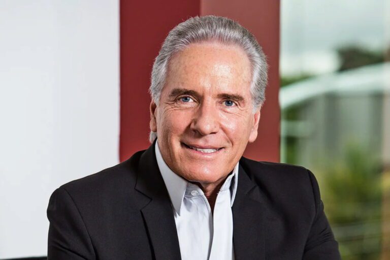 Retrato de Roberto Justus sorrindo, vestindo um terno preto, com fundo desfocado em um escritório moderno.