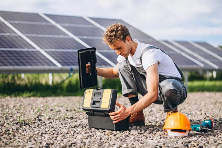 Técnico manuseando bateria solar em área externa, próximo a painéis solares, com ferramentas e capacete de segurança ao lado.