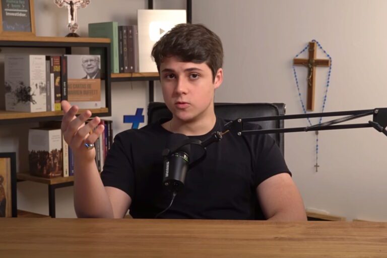 Felipe Molero, do Kids Investor, em um microfone em um ambiente de escritório, com estante de livros ao fundo e crucifixo na parede.