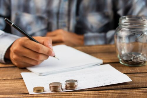 Homem escrevendo em caderno com moedas e jarro de vidro ao lado, representando o cálculo de valores em notas promissórias.