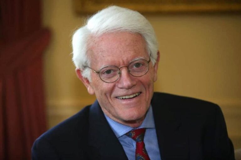Peter Lynch sorrindo, com cabelo branco e usando óculos, vestindo terno escuro e gravata vermelha.