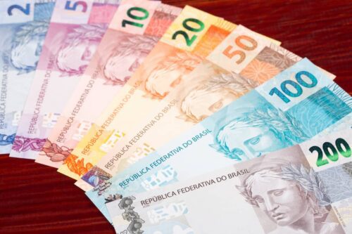 Cédulas de reais organizadas em leque sobre superfície de madeira, representando a diversidade da moeda fiduciária brasileira.