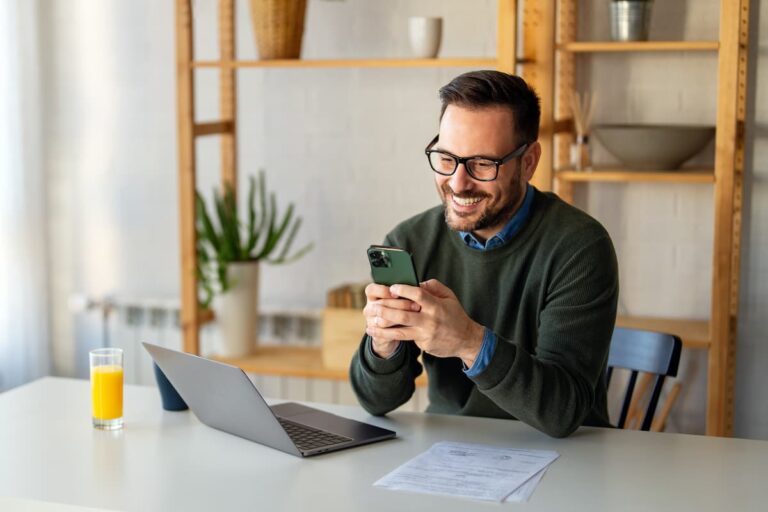 Homem sorrindo, usando celular em frente ao laptop com um copo de suco na mesa, simbolizando como ganhar dinheiro na internet de forma honesta e comprovada.