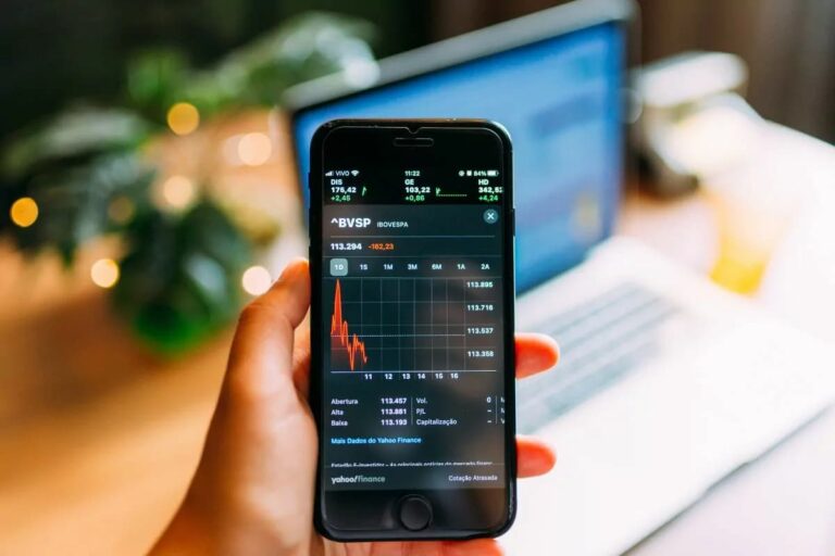 Smartphone exibe gráfico com queda no Ibovespa Futuro após declaração de Donald Trump; computador desfocado ao fundo.