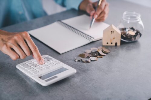 Pessoa fazendo cálculos em uma calculadora com moedas, caderno e uma casa em miniatura, simbolizando desconto nos FIIs.