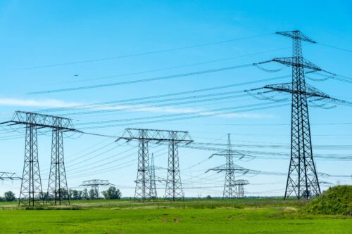 Torres de transmissão de energia elétrica em um campo verde sob céu azul, representando o Índice de Energia Elétrica (IEE na B3).