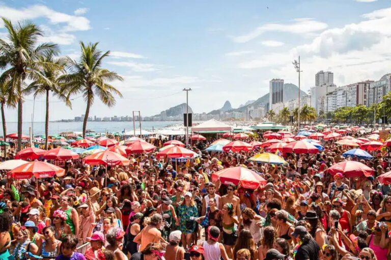 Multidão festeja nas areias de Copacabana sob guarda-sóis coloridos durante o Carnaval 2025. Hotéis do Rio de Janeiro lotados na folia.