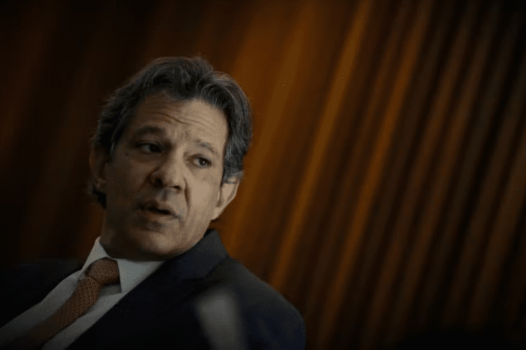 Haddad vestindo terno e gravata, falando em ambiente de escritório com fundo de cortinas de madeira, representando discussão sobre proposta do governo de alíquota fixa de IR de 17,5% para aplicações financeiras.