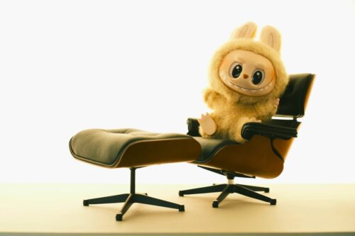 Labubu em roupagem felpuda sentado em poltrona de couro preto com apoio para os pés em fundo claro minimalista.