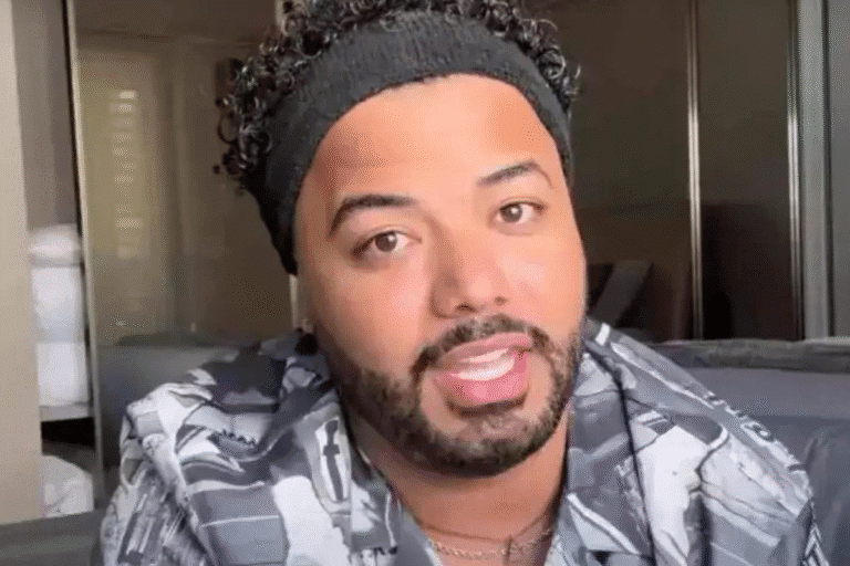 Hytalo Santos com cabelo cacheado usando faixa de cabeça e camisa de padrões geométricos, sorrindo para a câmera em ambiente interno.