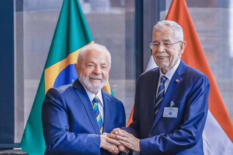 Duas pessoas em traje formal apertando as mãos em frente às bandeiras do Brasil e de outro país, simbolizando acordo ou cooperação internacional.