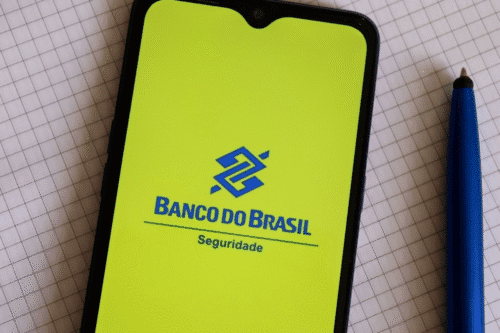 Imagem de um smartphone exibindo o aplicativo do Banco do Brasil na tela, com o logo azul e fundo amarelo, ao lado de uma caneta azul sobre papel quadriculado.