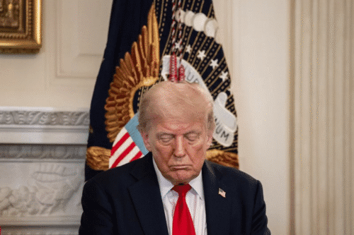 Imagem de Donald Trump com expressão séria, usando terno escuro e gravata vermelha, em um ambiente formal com bandeira e decoração ao fundo.