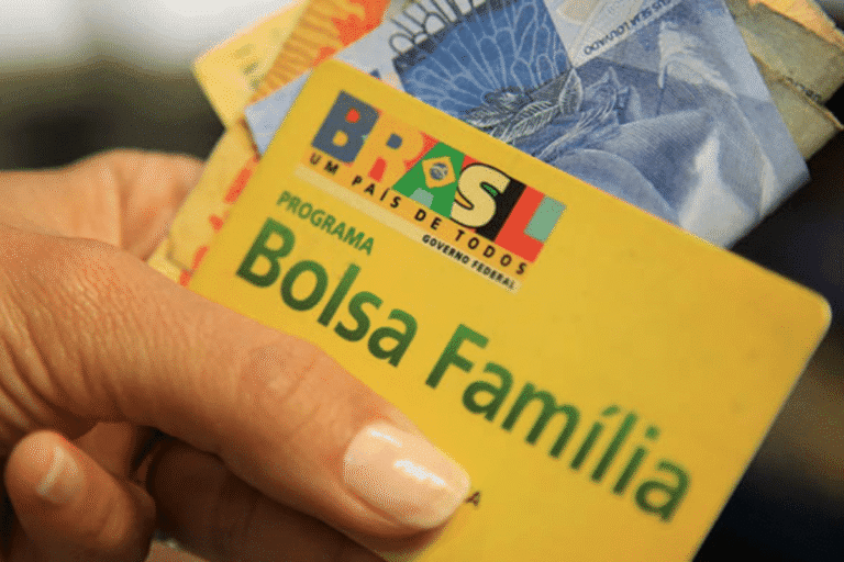 Imagem de uma mão segurando um cartão de benefício do Programa Bolsa Família com dinheiro do Brasil ao fundo, destacando a importância do programa social para famílias brasileiras.