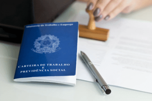 Imagem de uma carteira de trabalho e previdência social sobre uma mesa, com uma caneta e uma mão usando um carimbo ao fundo, representando documentos trabalhistas e previdenciários.