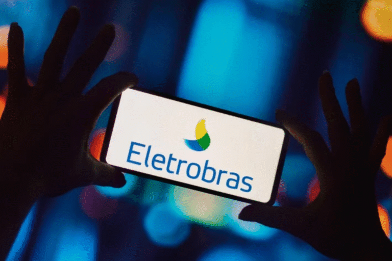 Pessoa segurando um celular exibindo o logotipo da Eletrobras, empresa de energia elétrica no Brasil, com fundo de luzes coloridas e ambiente noturno.