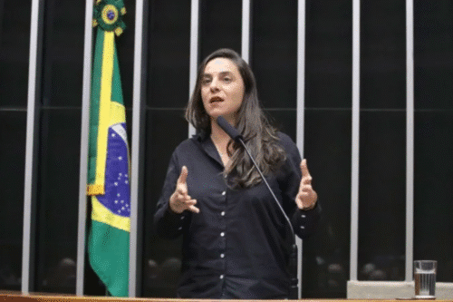 Mulher falando em público ao lado da bandeira do Brasil, em ambiente de tribunal ou conferência, vestida com camisa preta e gesticulando.
