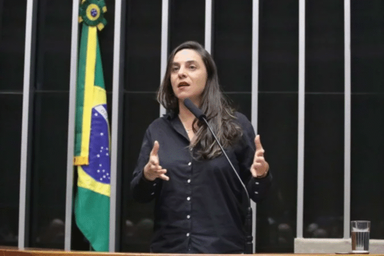 Mulher falando em público ao lado da bandeira do Brasil, em ambiente de tribunal ou conferência, vestida com camisa preta e gesticulando.