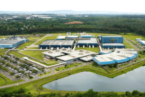 Visão aérea de um grande parque tecnológico com centros de pesquisa, áreas de estacionamento e uma lagoa ao redor, em um ambiente com zona rural ao fundo.