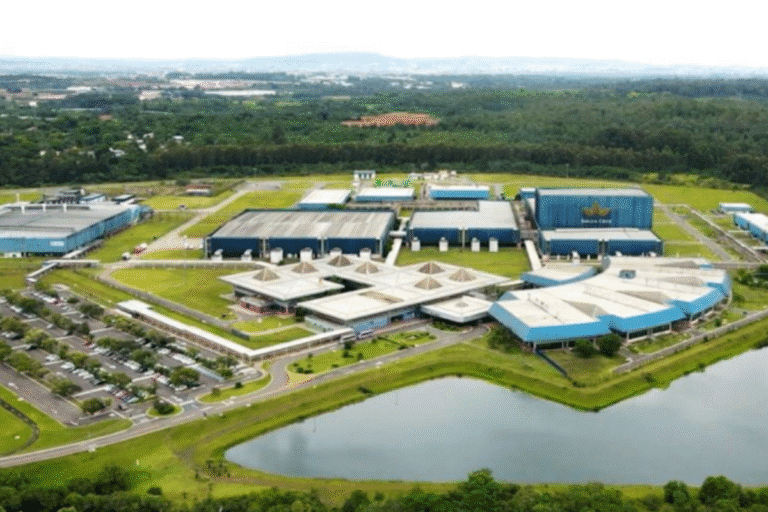 Visão aérea de um grande parque tecnológico com centros de pesquisa, áreas de estacionamento e uma lagoa ao redor, em um ambiente com zona rural ao fundo.