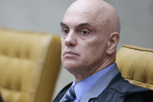Alexandre de Moraes em tribunal, com expressão séria, usando beca preta e gravata, sentado em uma cadeira de tribunal
