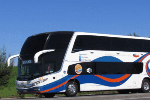 Ônibus de turismo branco com detalhes em azul e laranja, rodando em estrada sob céu claro, pronto para viagens de lazer.